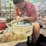 Person shaping clay on a pottery wheel in a bright studio with shelves and tools in the background