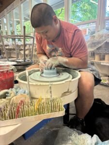 Person shaping clay on a pottery wheel in a bright studio with shelves and tools in the background