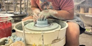 Person shaping clay on a pottery wheel in a bright studio with shelves and tools in the background.