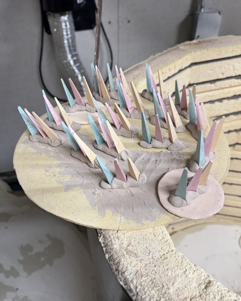 Colorful pastel triangular sculptures inserted into soft clay bases on a dusty workshop shelf arranged in rows art project in progress
