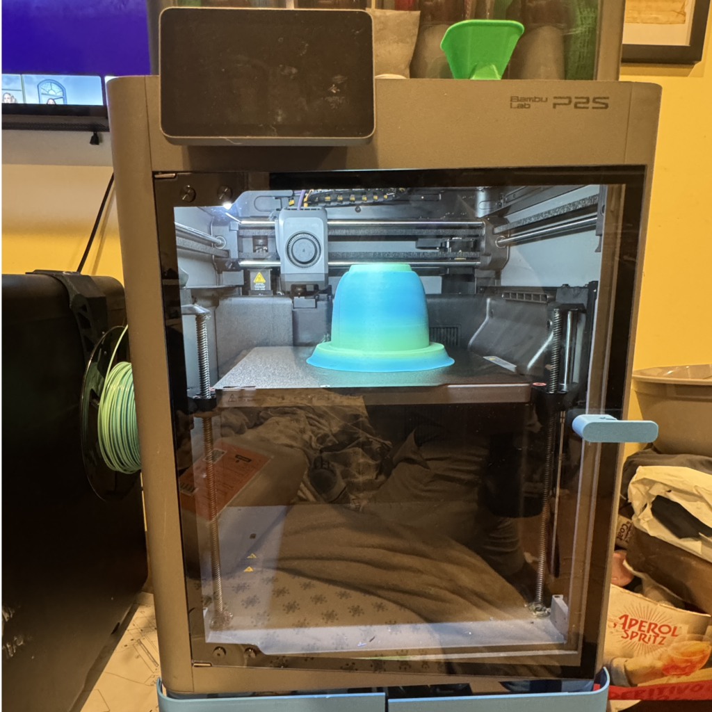 3d printer inside a cabinet with a blue filament spool on the left and a turquoise object on the print bed